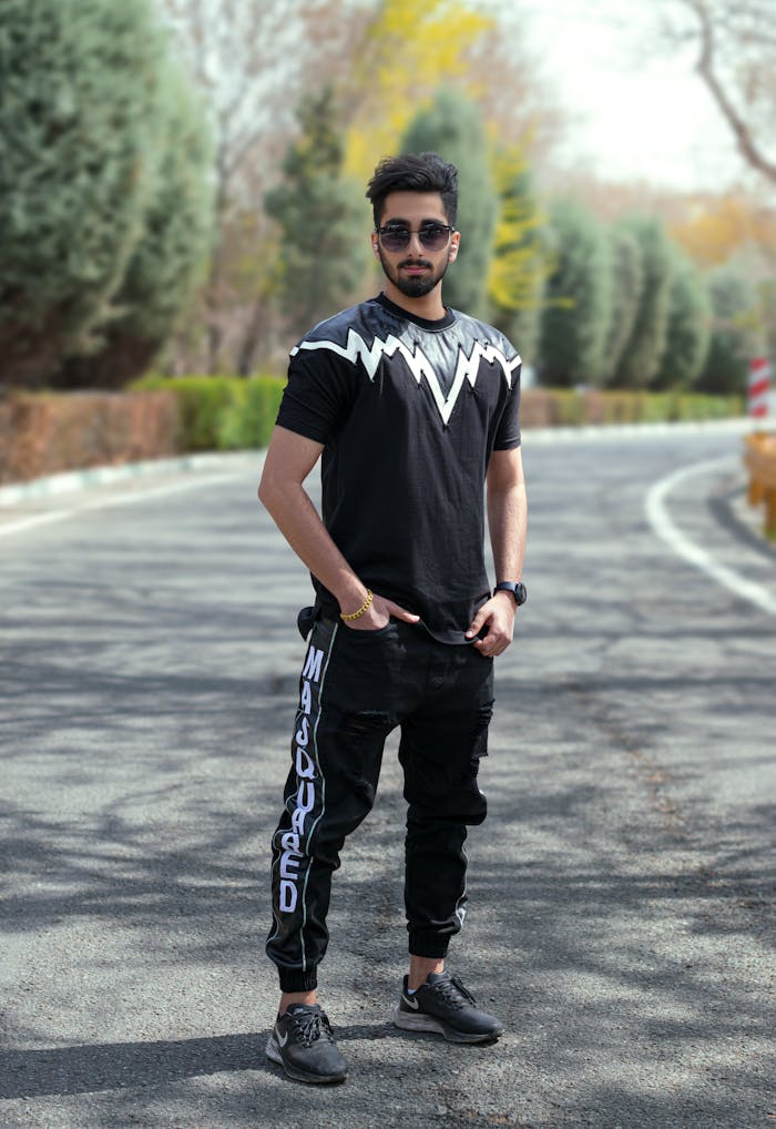 A fashionable man wearing sunglasses and a black outfit poses confidently on a city street.