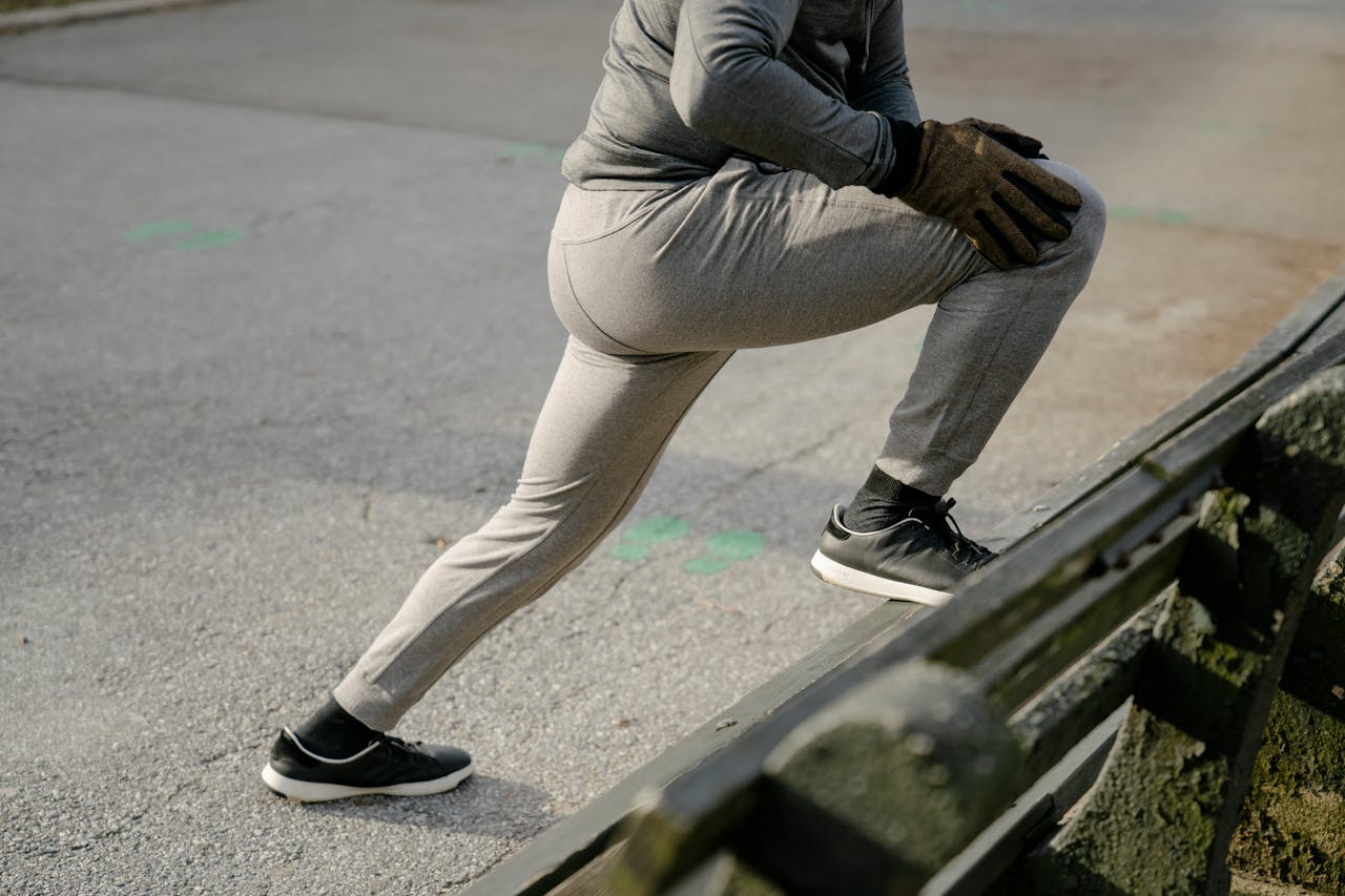 Crop anonymous male athlete wearing warm activewear stretching legs before training on asphalt road in city park in daylight